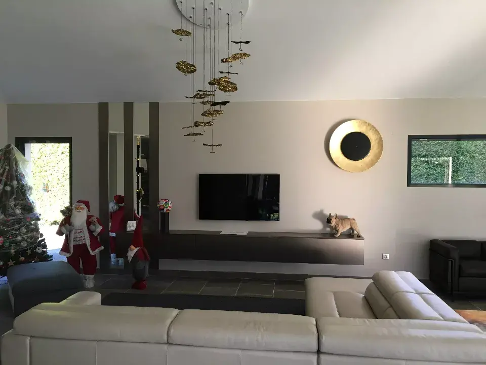 Grand salon avec magnifique luminaires couleur doree. A gauche un sapin et un pere noel. Au centre un meuble mural blanc sur lequel repose une television et une figurine de chien (bulldog). A droite une assiette doree accrochee au mur / decoration par ViolaDeco, Iana Viola