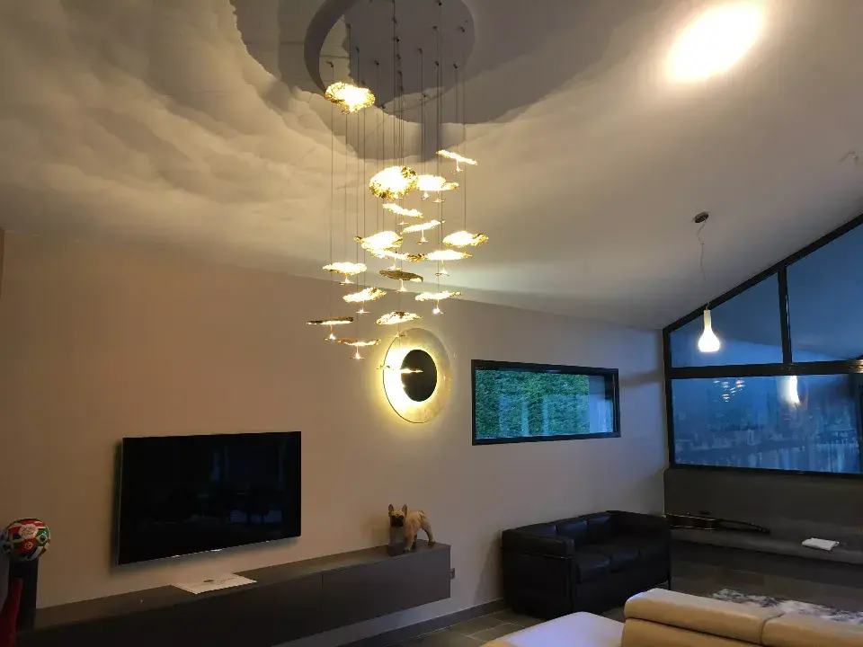 Grand salon avec un magnifique plafonnier central creant des effets de lumieres. Au centre, un meuble mural blanc sur lequel repose une television et une figurine de chien (bulldog). A droite un canape noir et un luminaire sur le mur en forme d'assiette et de couleur doree / decoration par ViolaDeco, Iana Viola